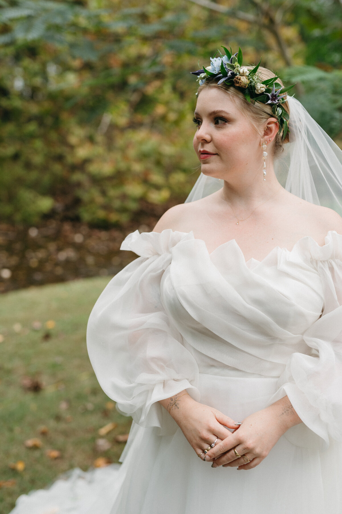Bride posing outdoors in an ethereal off-the-shoulder organza gown with voluminous sleeves, wearing a botanical floral crown and holding a seasonal garden bouquet created by a wedding florist in Arkansas.