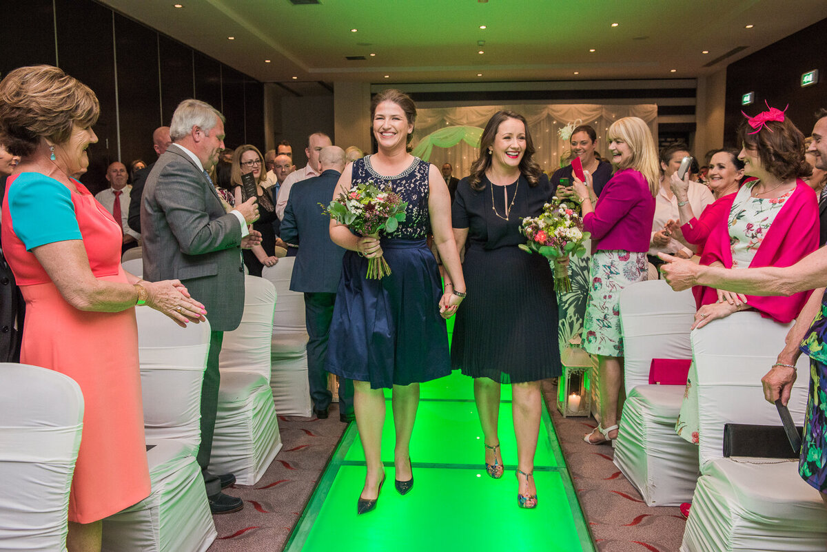 Two brides walking up a neon green aisle wearing navy dresses