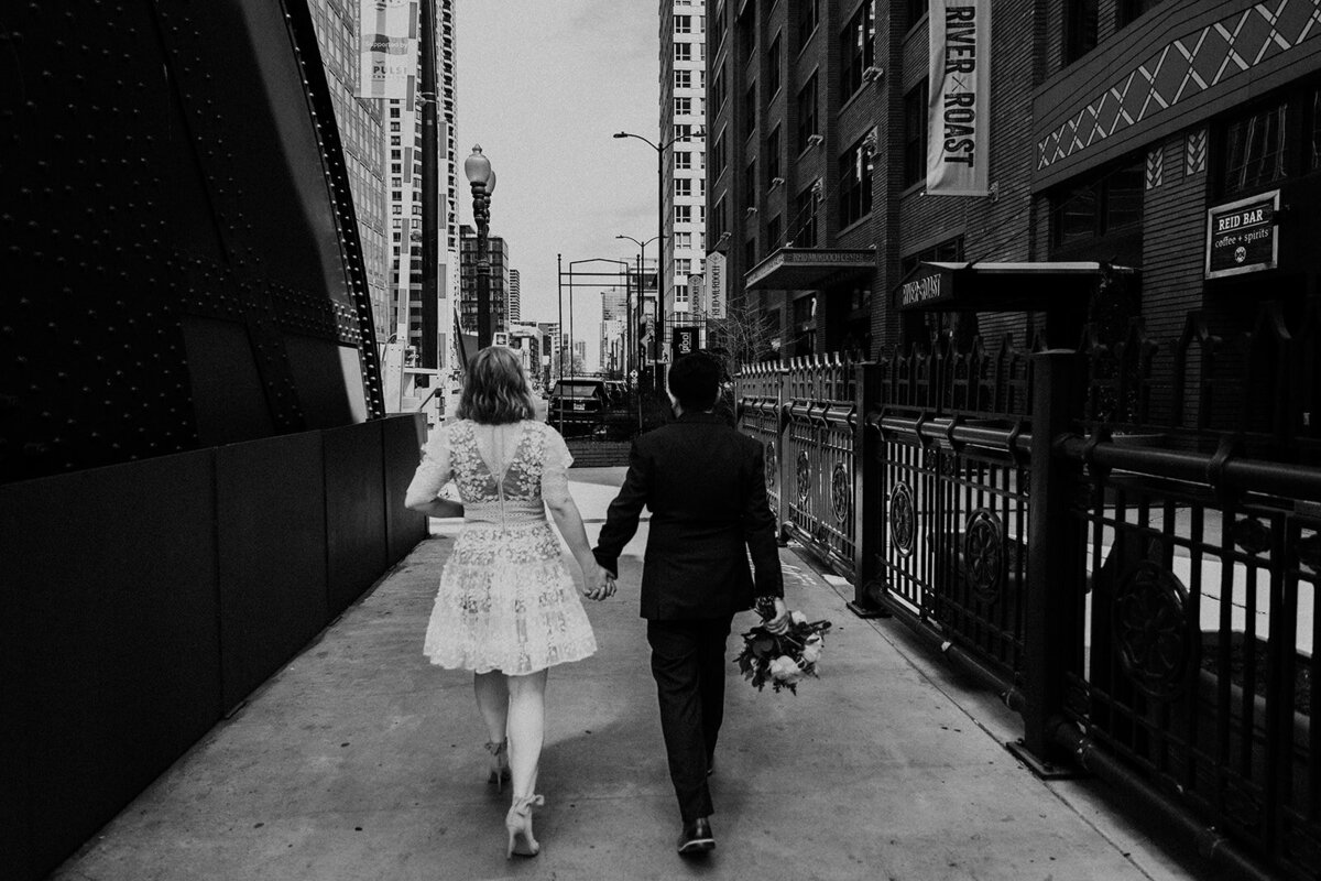 Elopement couple walking away from camera across the Chicago River on a bridge. They're holding hands and the groom is carrying her bouquet.