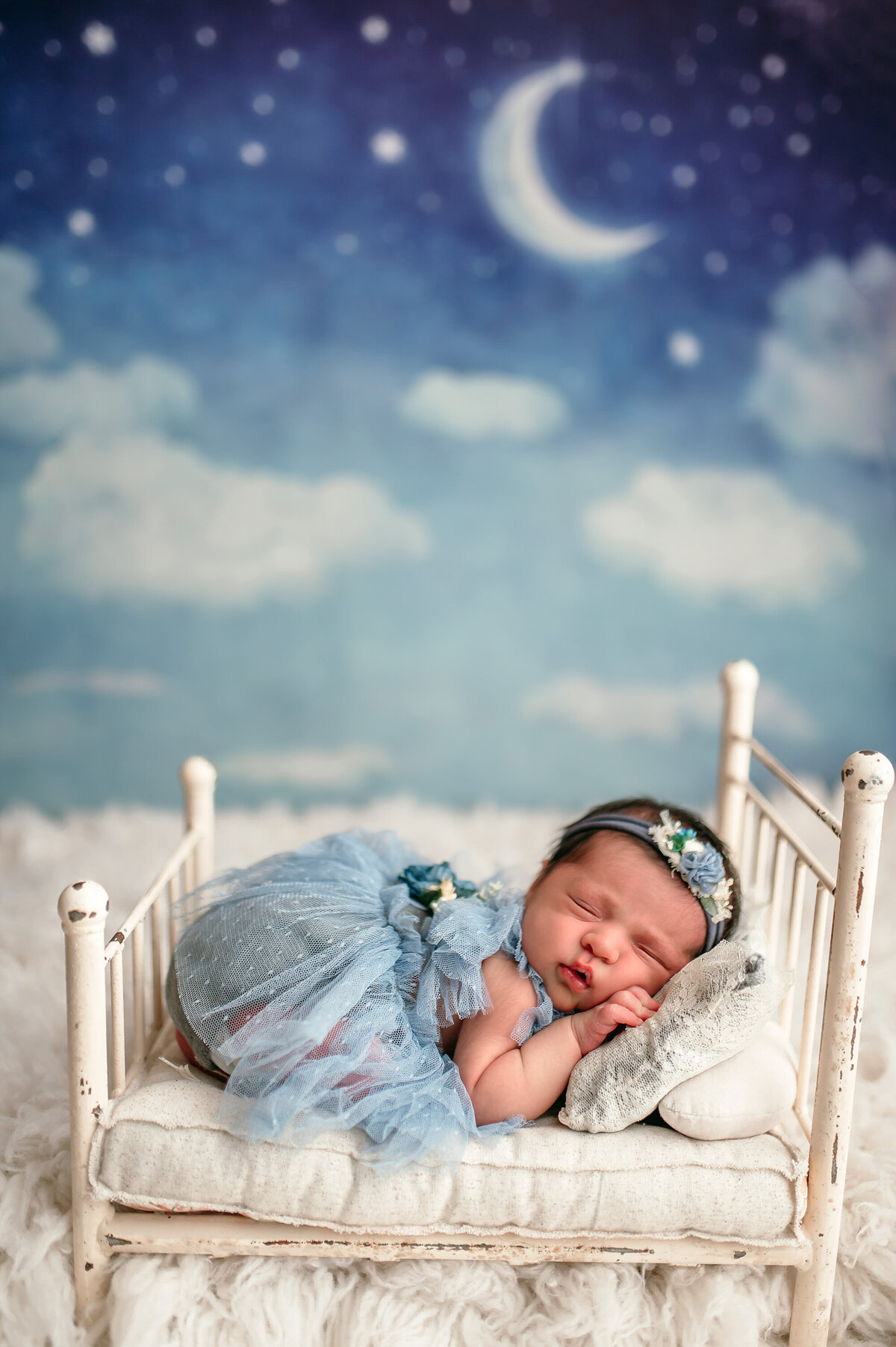 Night time themed newborn portrait of a baby girl in a blue dress asleep on a tiny doll bed in front of a moon and star backdrop.