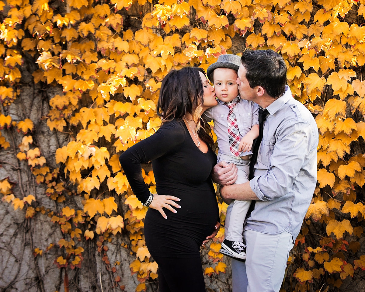 Mom and dad holding toddler boy and kissing his cheeks while mom is holding her pregnant belly in front of yellow autumn leaves.