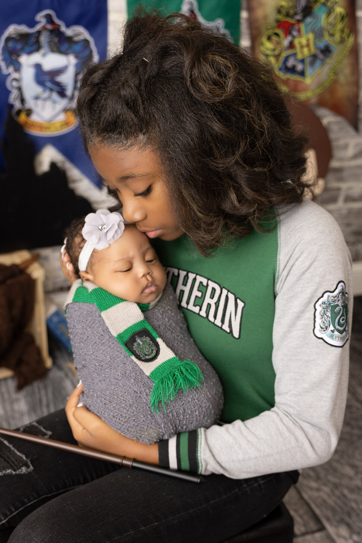 A young girl wearing a green and grey Hogwarts-themed sweater lovingly holds and kisses a sleeping baby wrapped in a Slytherin scarf. Banners of different Hogwarts houses hang in the background, beautifully captured by a Jacksonville newborn photographer.