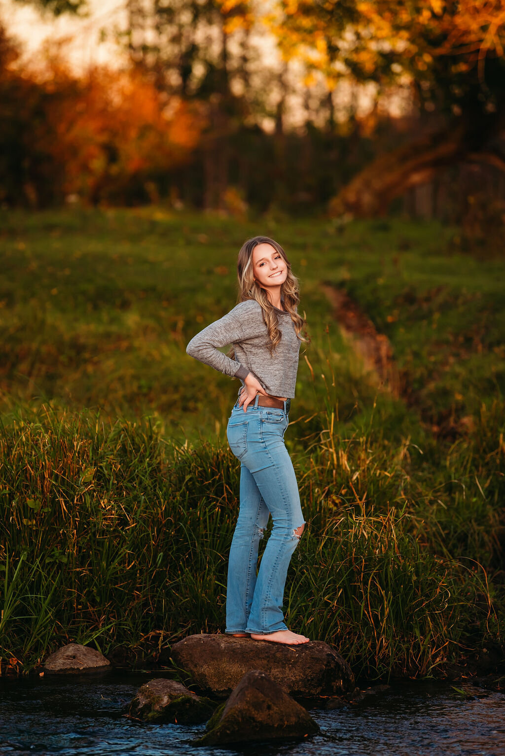 A young woman standing in a river at sunset, with warm light illuminating her joyful expression and flowing hair, captured by a Bemidiji, Minnesota fine art senior photographer
