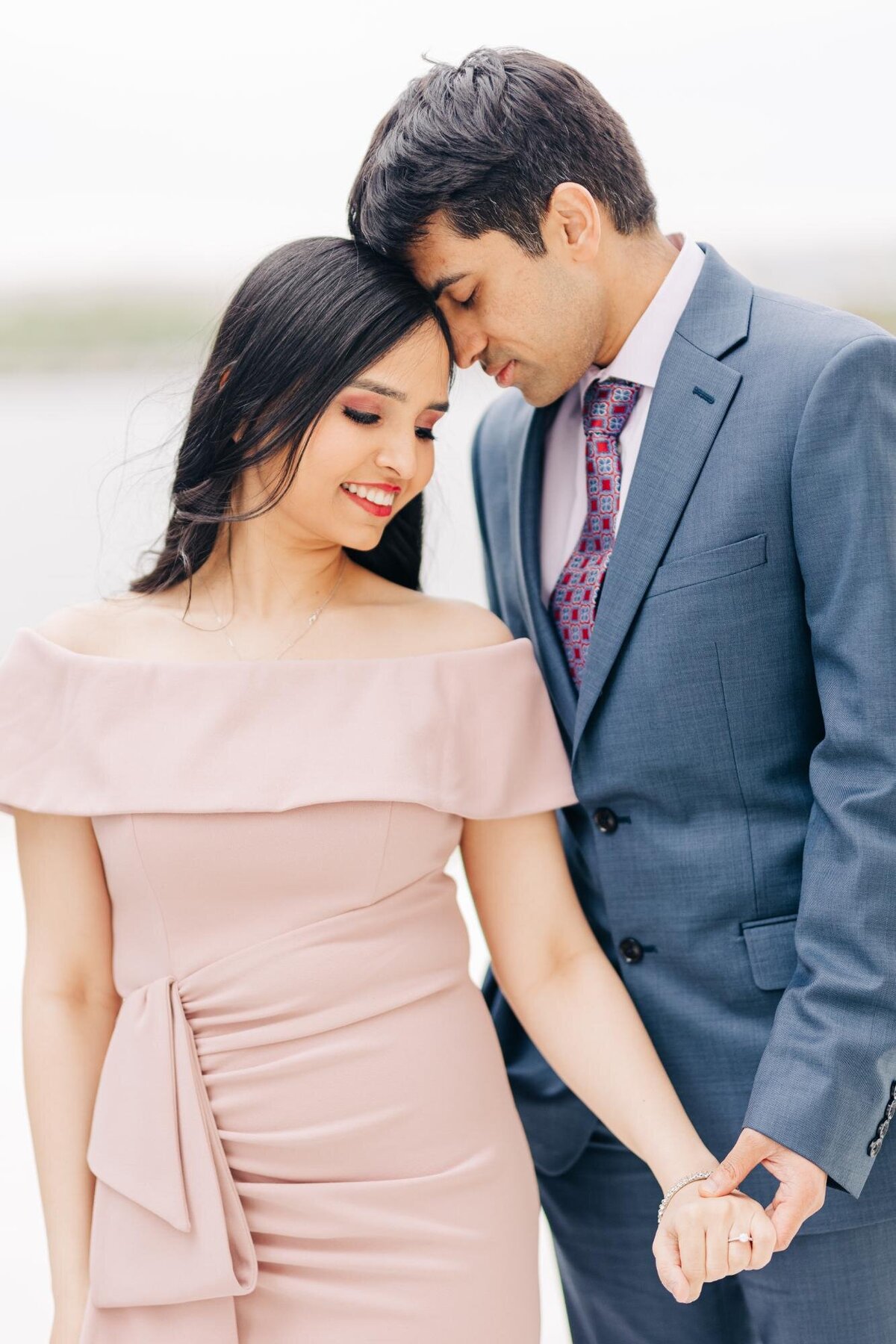 A couple dressed formally, the man in a grey suit and the woman in a pink off-shoulder dress, stand close together, smiling gently and holding hands.
