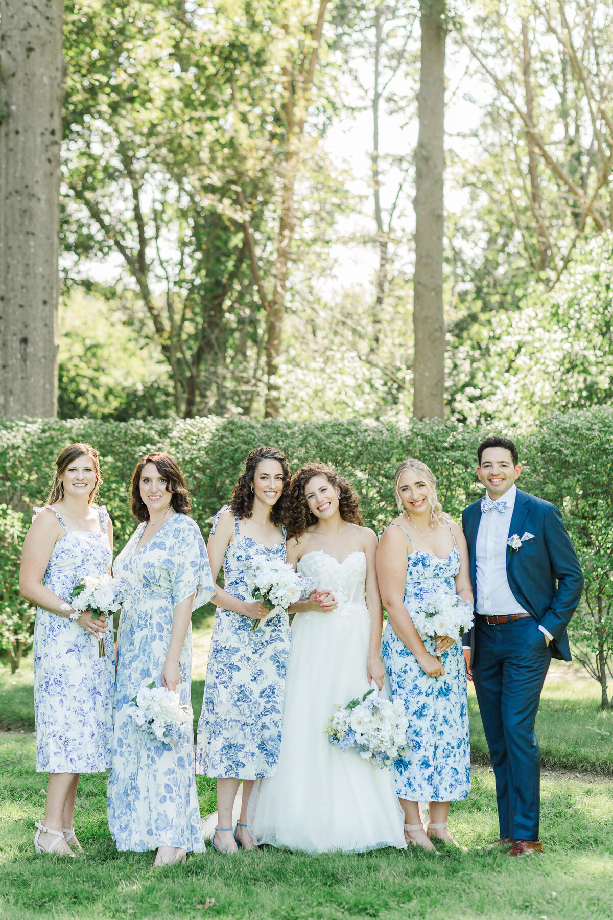 A bride is standing on the grounds of Glen Magna Farms in Danvers, MA with her bridal party. All of them are smiling at the camera and standing casually. The bridesmaids are in various blue and white patterned dresses. Captured by MA wedding photographer Lia Rose Weddings