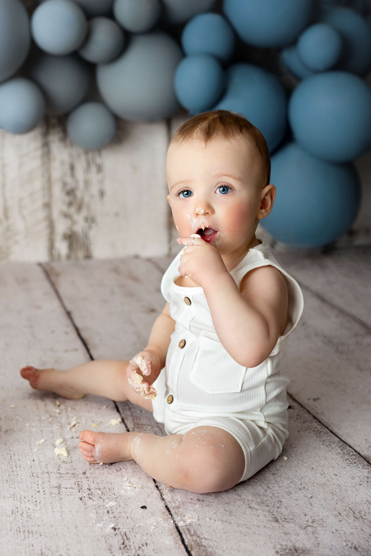 one year old boy sitting to the side putting cake in his mount with blue balloons behind him