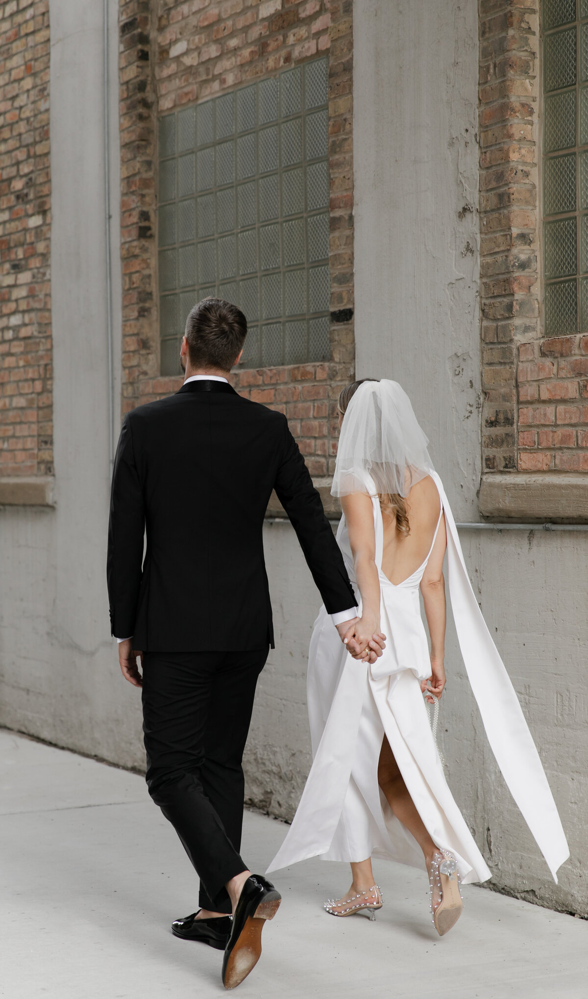 Bride in elegant low-back gown holds hands with groom as they are pictured from behind outdoors at Chicago wedding ceremony.