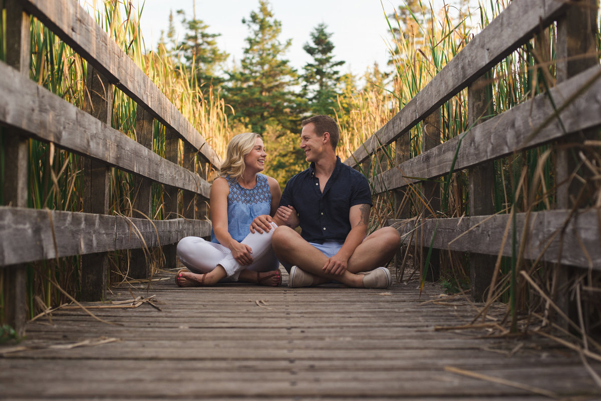 couple sitting cross-legged on wooden bridge next to one another laughing