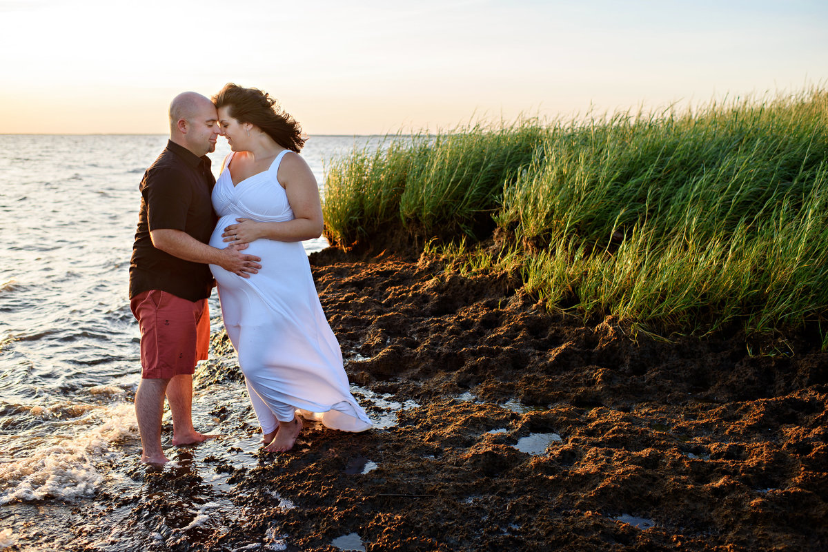 A pregnant couple stand in the water at the beach and hold each other.