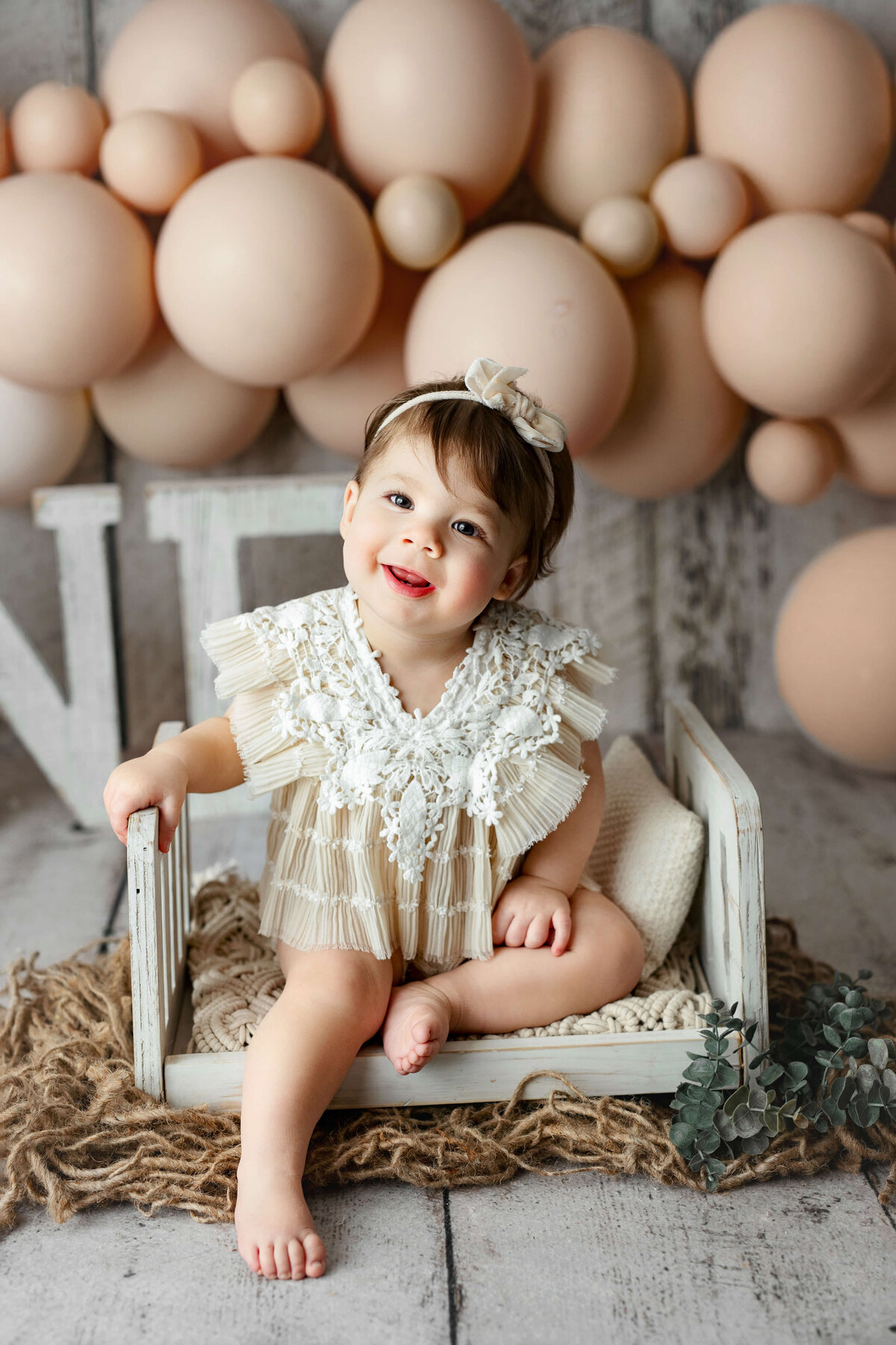 toddler in a lacey white outfit and hair bow sitting on a white wood during a milestone baby session