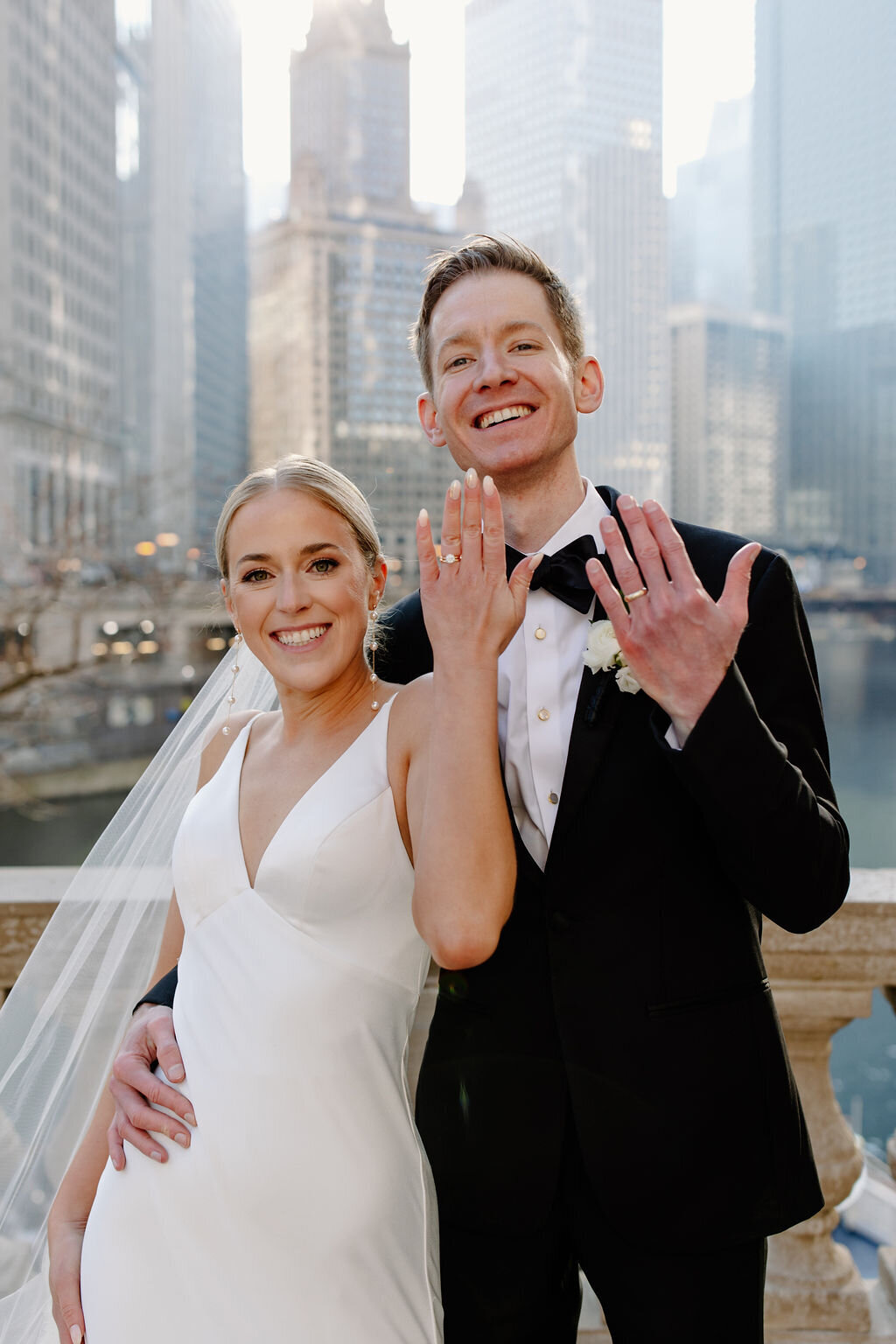 Bride and groom smile to camera and hold up hands to indicate their wedding rings while overlooking Chicago skyline