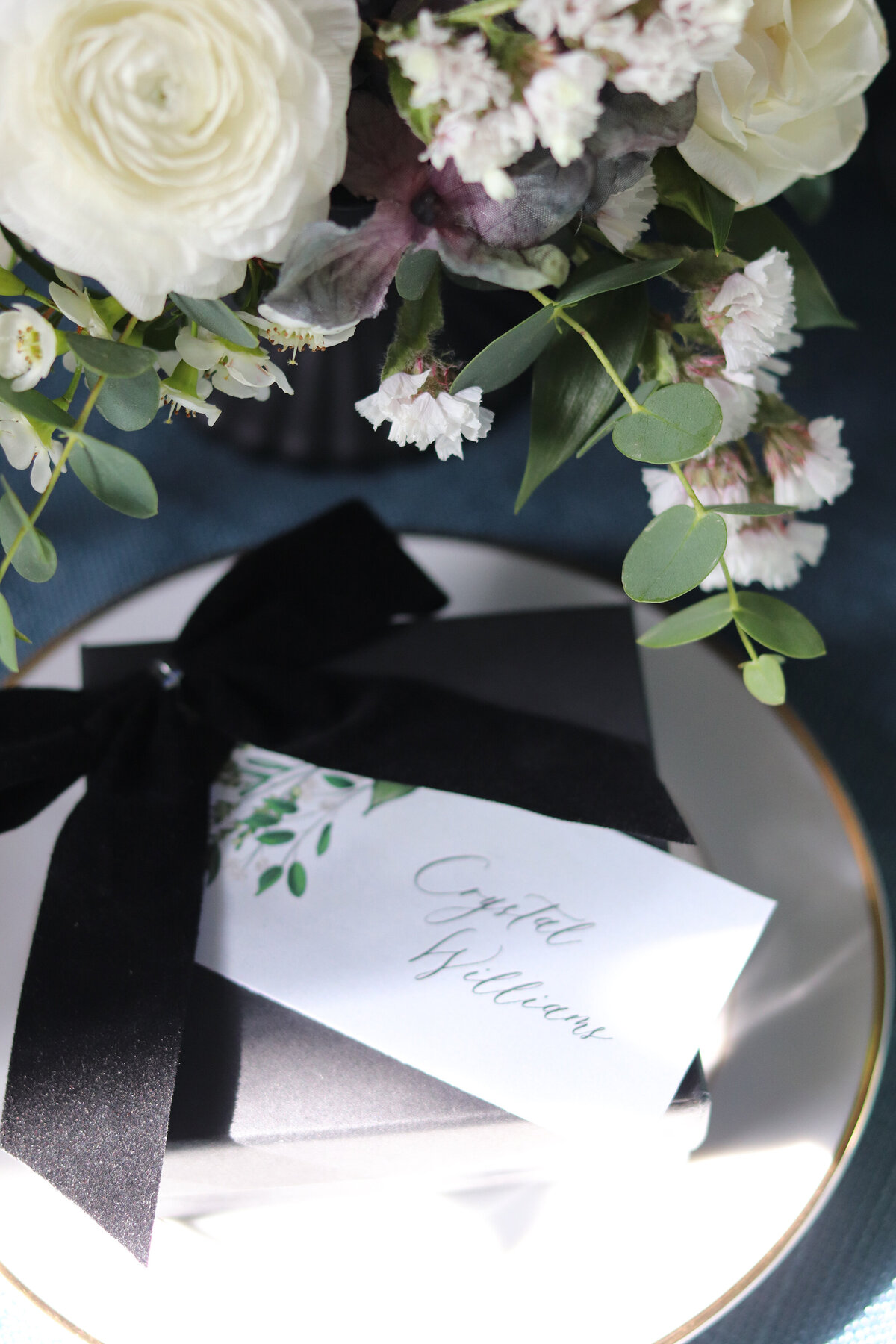 A wedding favor with a black velvet ribbon and a floral name tag sitting on a small plate next to a flower arrangement