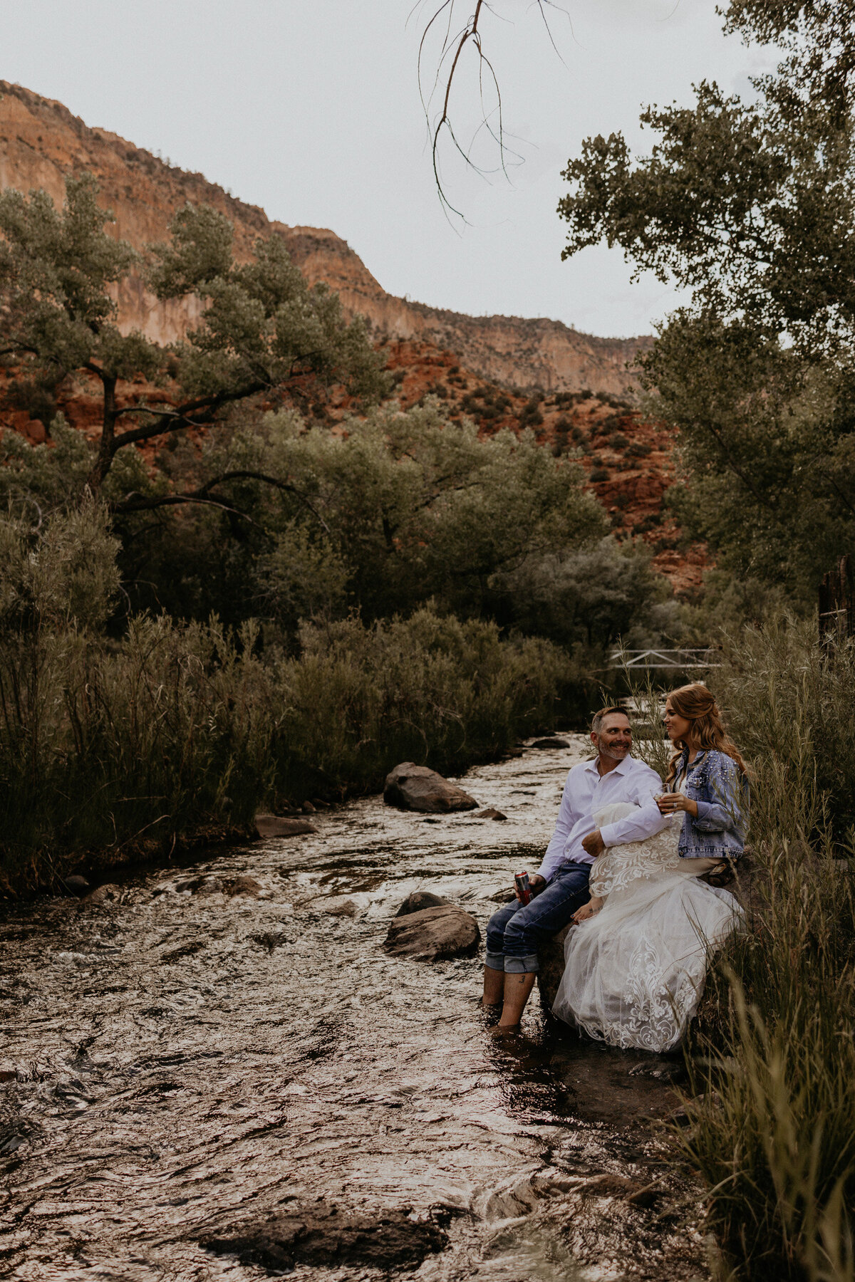 newlyweds enjoying a drink together sitting along a creek after their  elopement