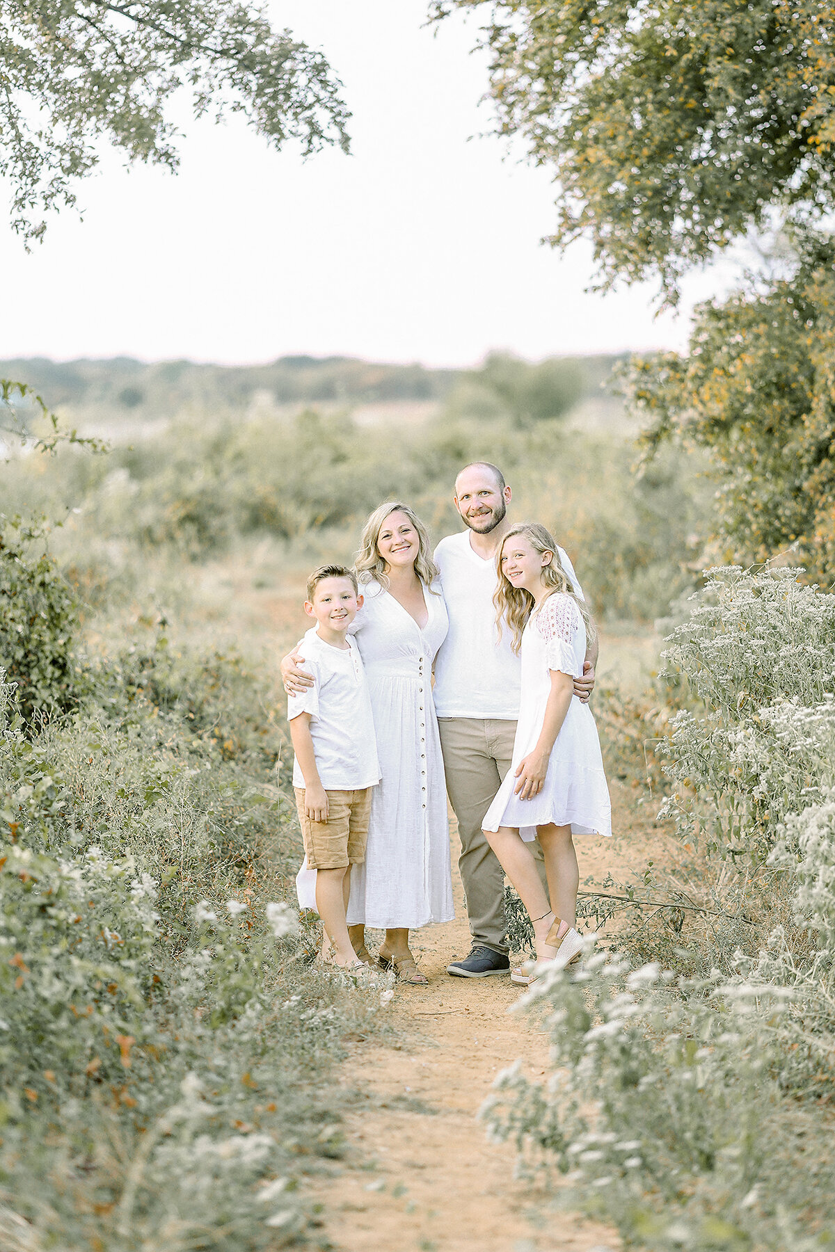 A family photo taken while they are standing together holding hands in the middle of a path by a beautiful field at a local Dallas Fort Worth park captured by their favorite Dallas Family Photographer.
