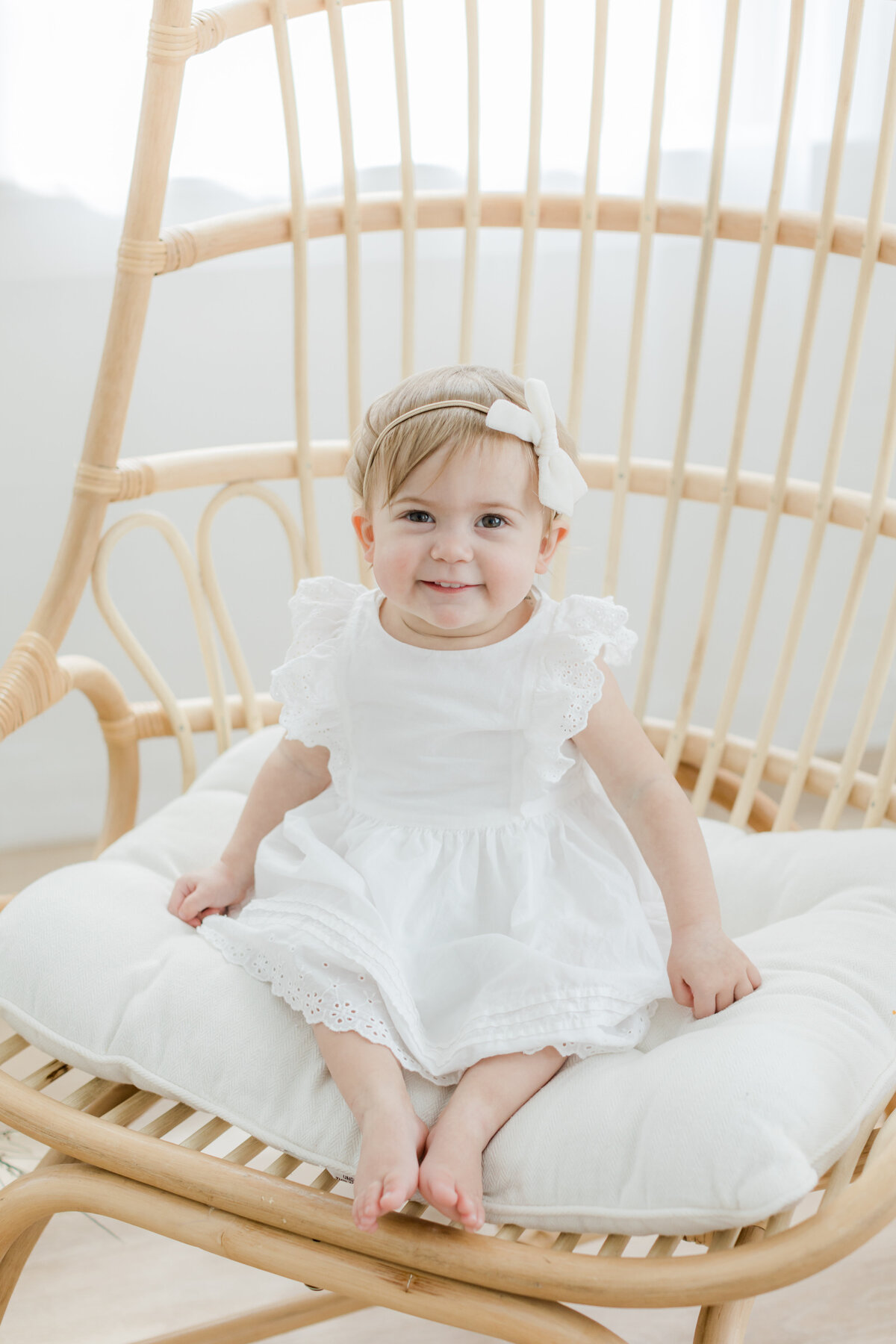 toddler girl wearing a white dress with a bow in her hair sitting on a rattan chair photographed by Philadelphia Portrait Photographer Tara Federico