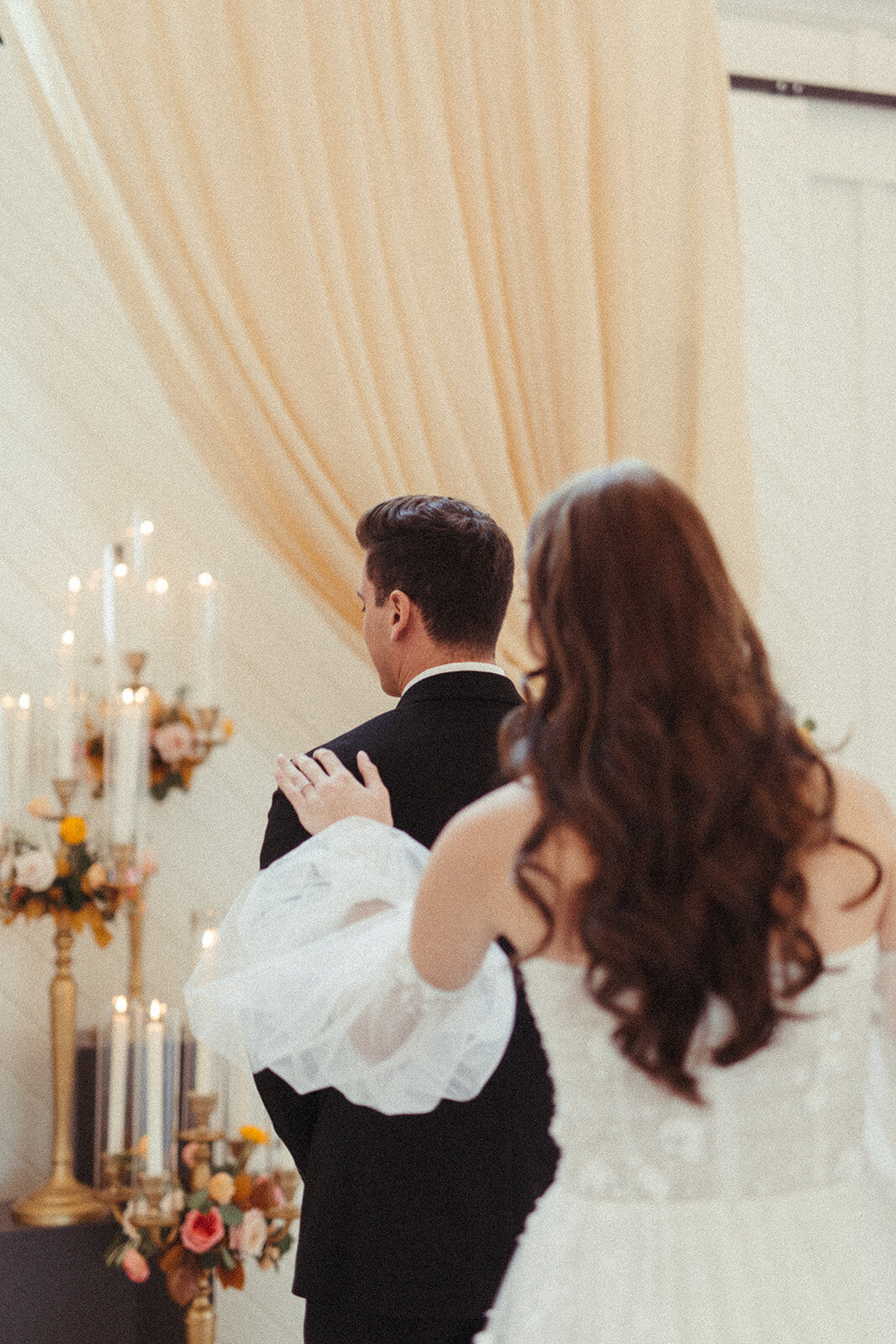Bride and groom wearing a white wedding gown and black tuxedo stand by lit candlesticks and peach-colored curtains.