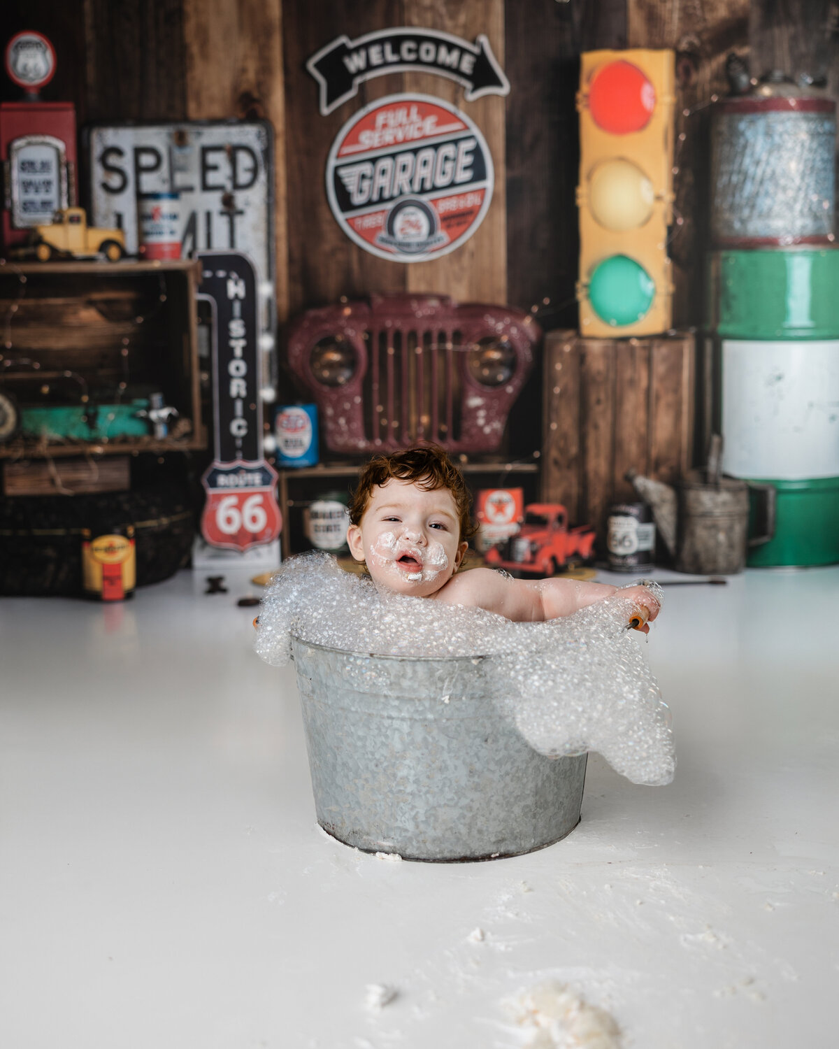 Baby boy with curly hair and a frosting-covered face sitting in a sudsy metal tub in a vintage automotive backdrop.
Photograph by Yucaipa cake smash photographer Kaitlyn Dawn Photography.