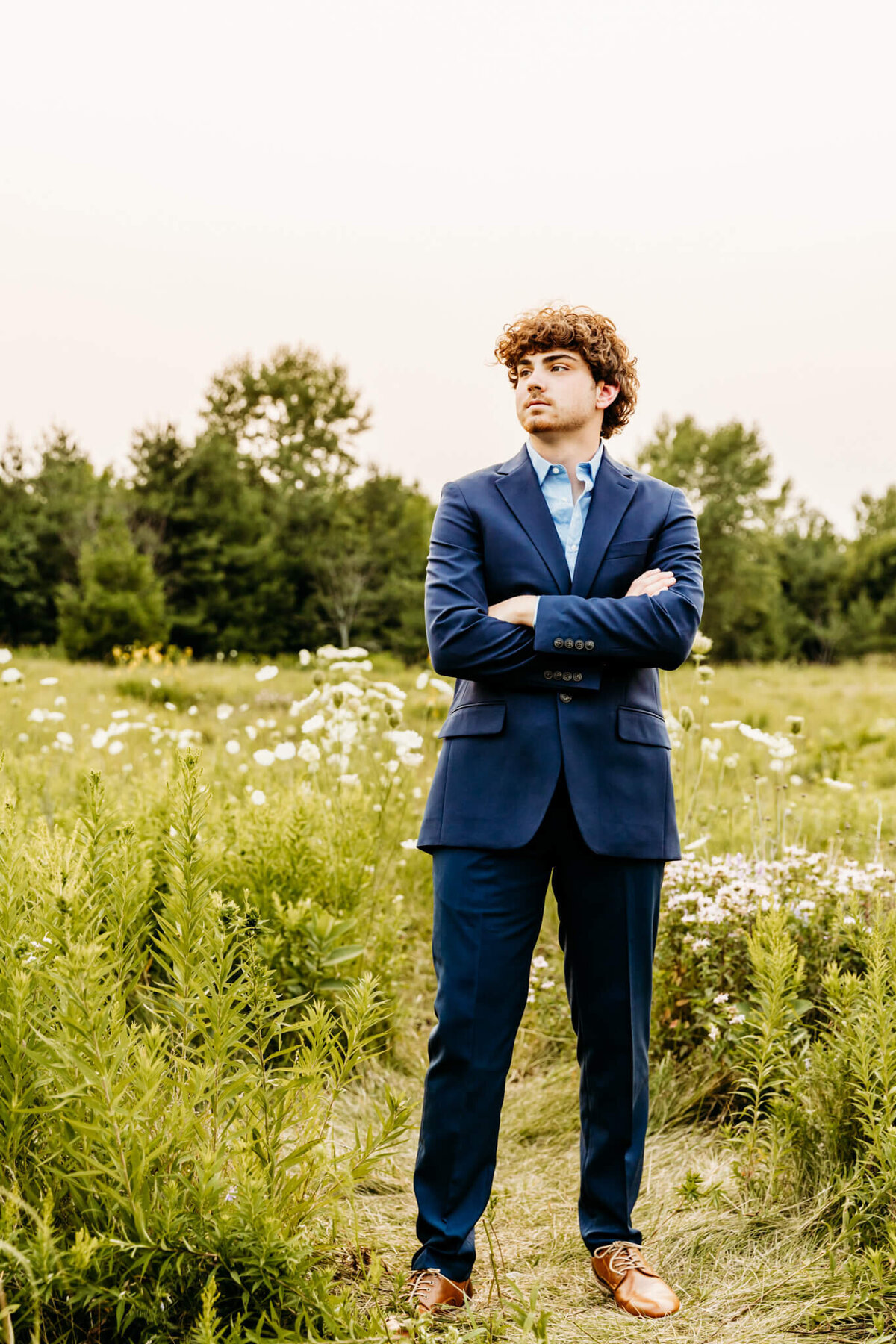 young man surrounded by wildflowers in a blue suit crossing his arms and looking out