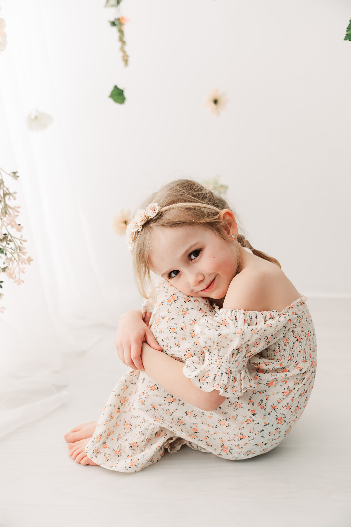 A little girl in a floral dress sitting on the floor, tilting her head charmingly with a gentle smile, in a bright room adorned with hanging flowers. Taken by Andover Newborn Photographer, Fig and Olive Photography.