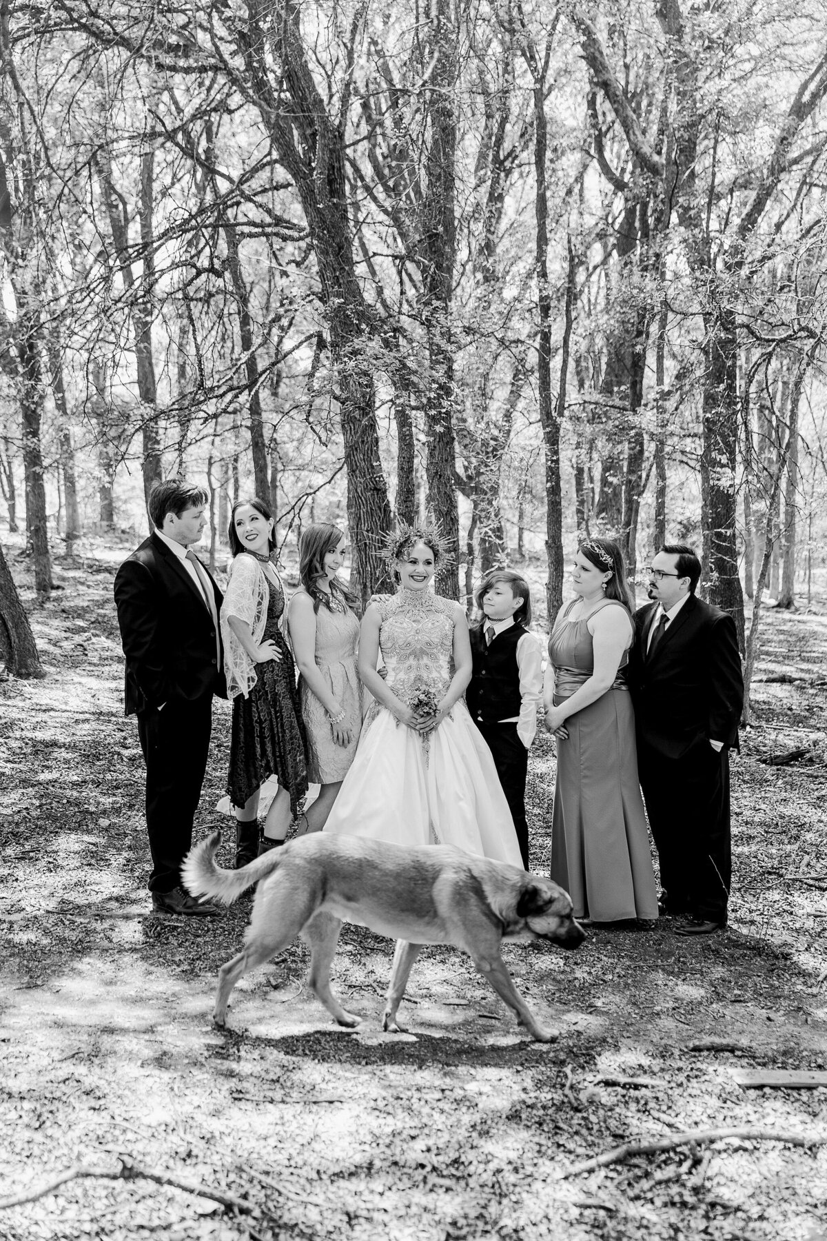 A black and white portrait of a bride, her dog, and her wedding party as posing together in the woods on her wedding day in Fort Worth, Texas. The pose is very casual with some people smiling, other looking at each other, looking at the bride, etc. The bride stands in the center and is wearing an elaborate, very intricate dress and crown while holding a bouquet. The wedding party is wearing varying dresses and suits.