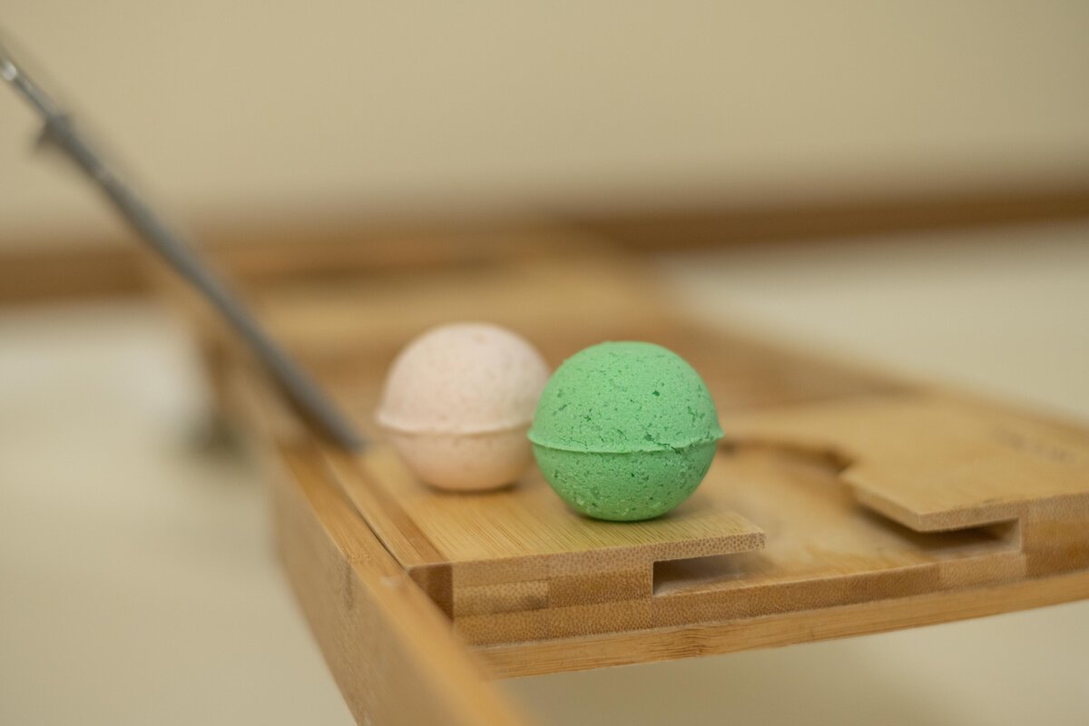Two bath bombs, one pink and one green, placed on a wooden tray next to a metallic handle.