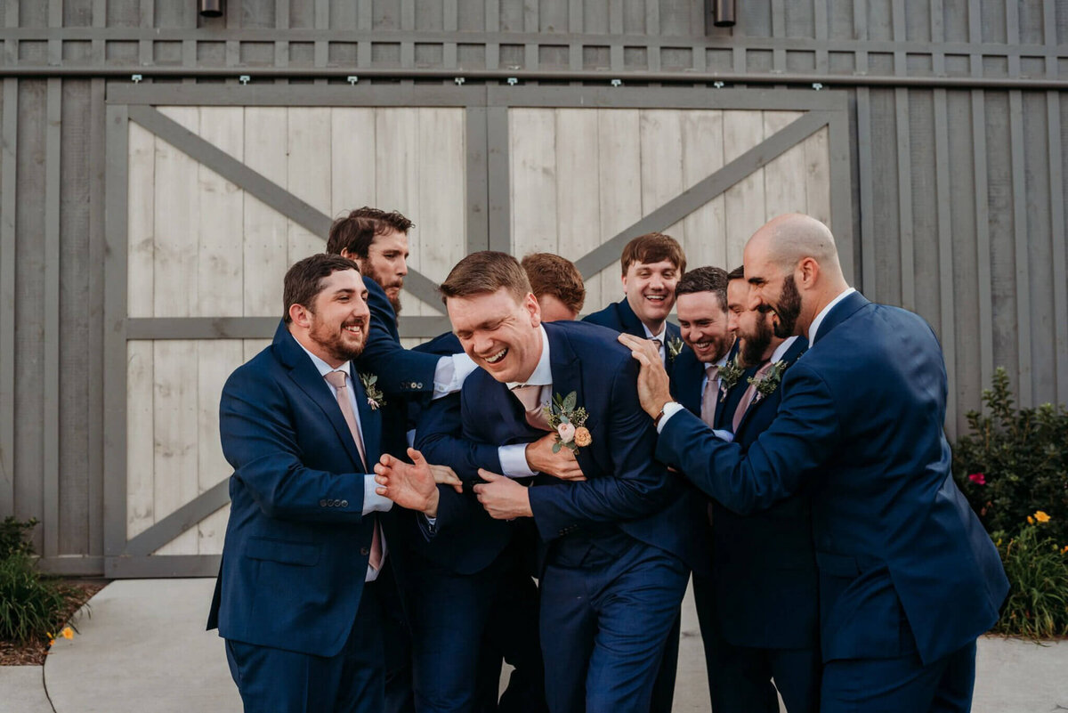 Photo of a groom and his groomsmen wearing navy suits and laughing in a huddle in front of gray barn doors