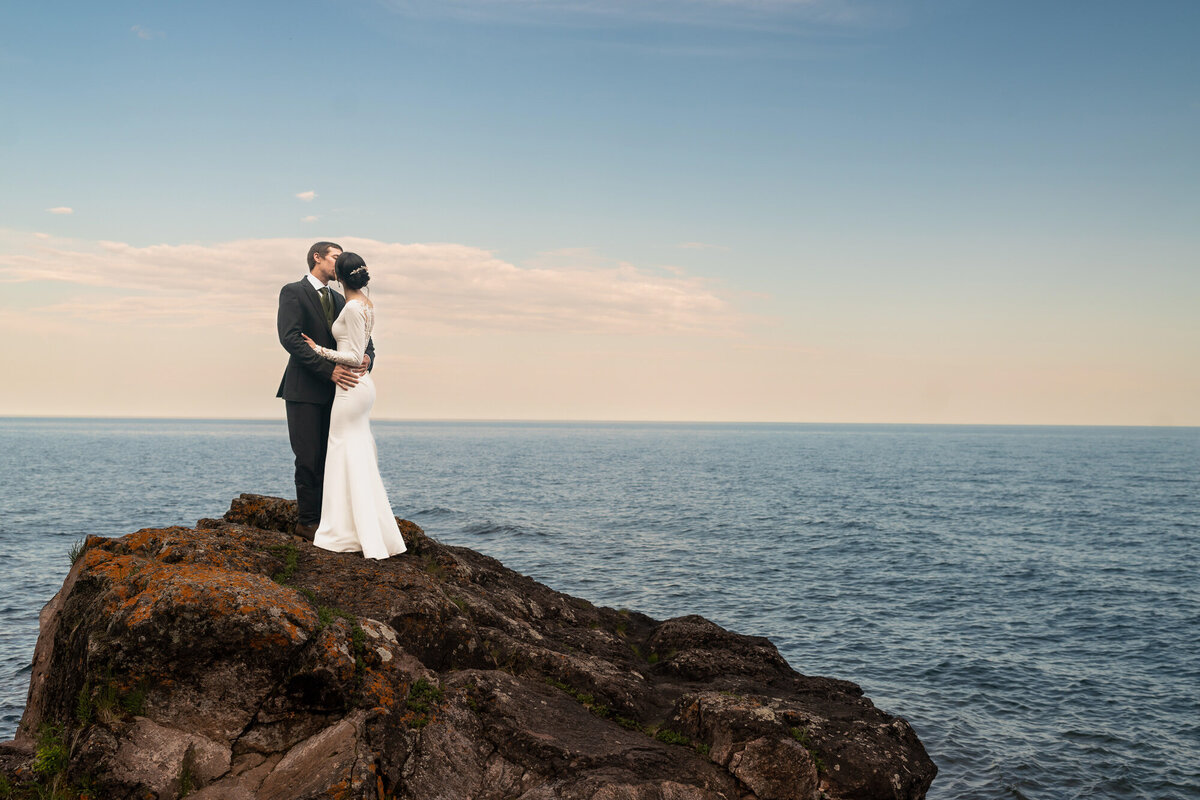 Bride and groom kiss on a rock at sunset in front of Lake Superior.