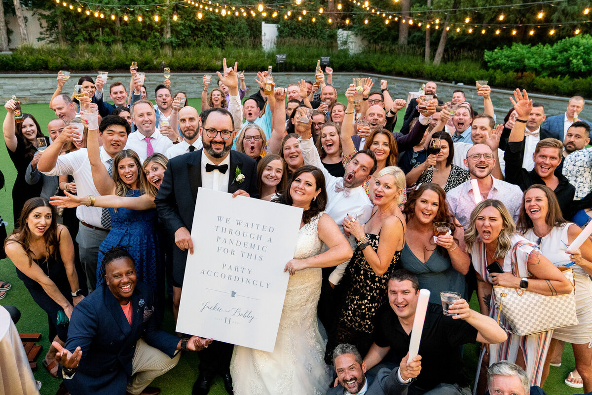 Bride and groom hold sign with guests at wedding reception.