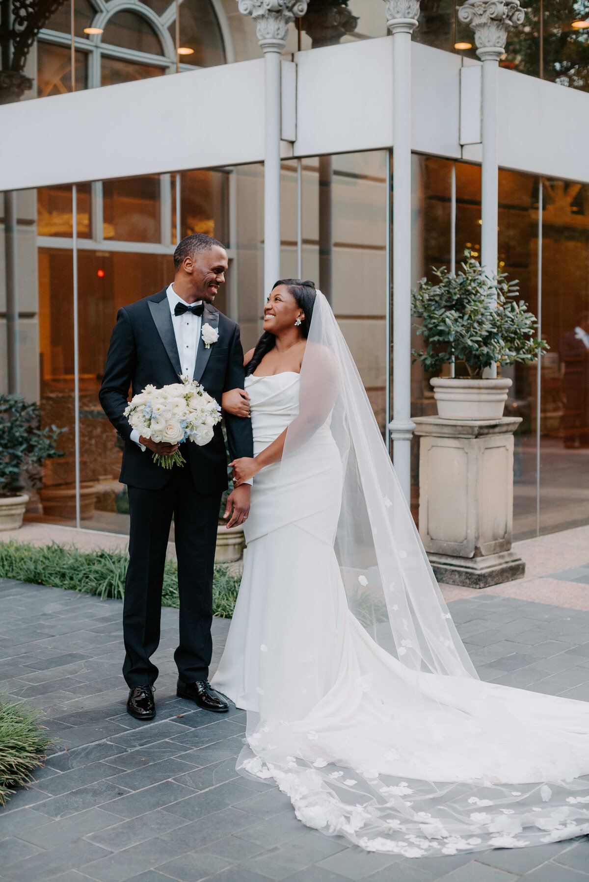A joyful couple stands in an elegant outdoor setting. The bride wears a flowing white gown and veil, holding the groom's arm. He wears a black tuxedo and holds a bouquet.