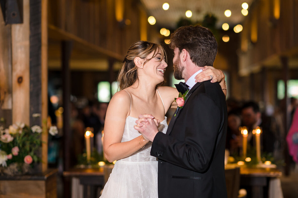 A bride and groom dancing intimately, surrounded by guests and candlelight in a wooden barn reception.