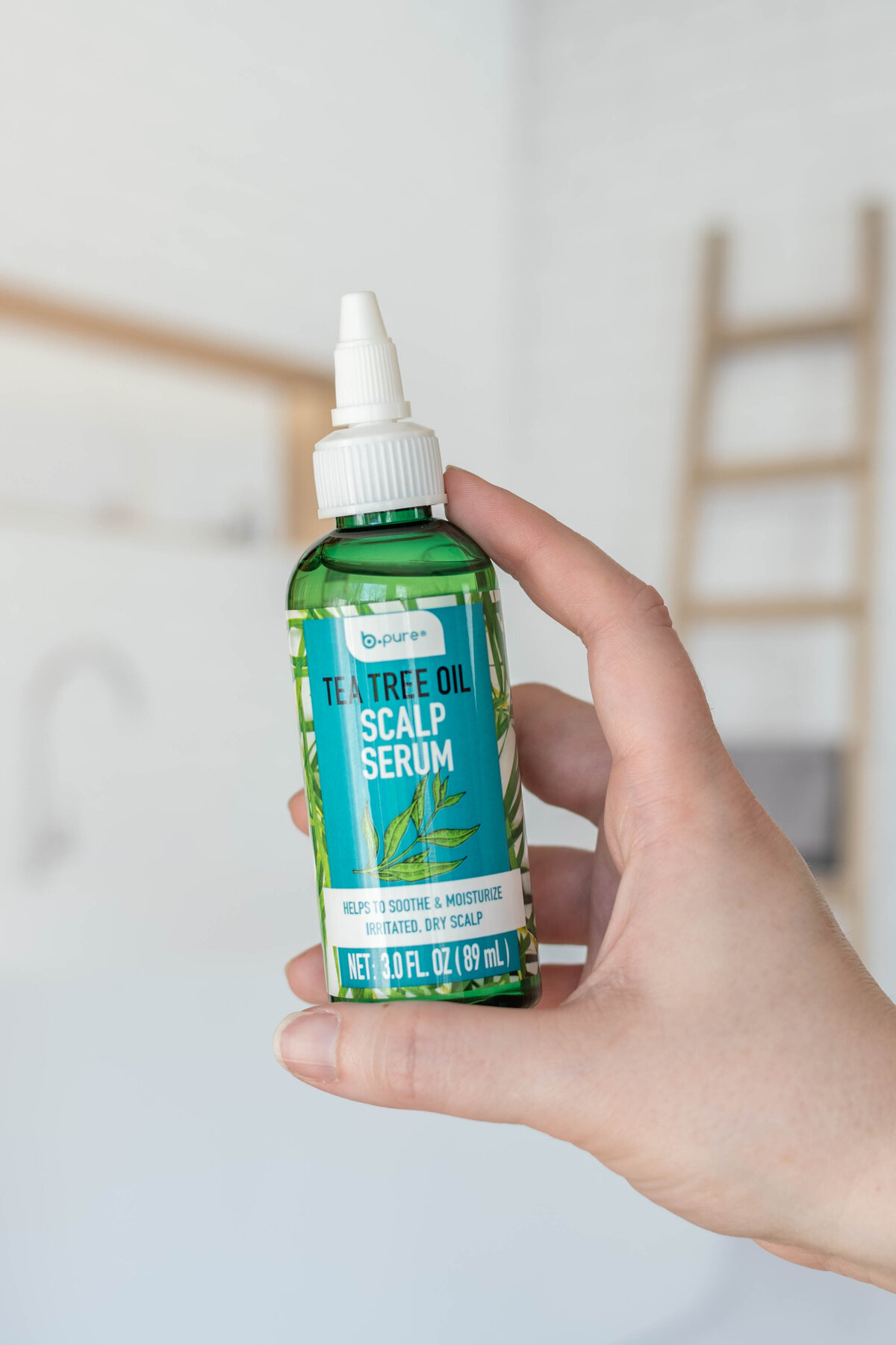A person holding a bottle of be-pure tea tree oil scalp serum in a bright, airy room.