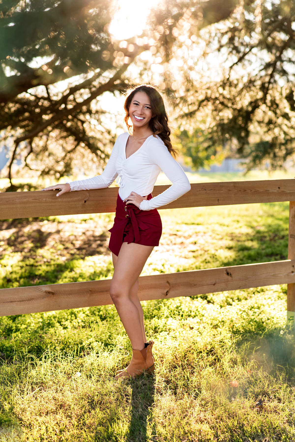 RVA high school senior girl stands against split rail fence at Maymont Park for her senior pictures.. Lovely backlight streams in through pine trees behind her.