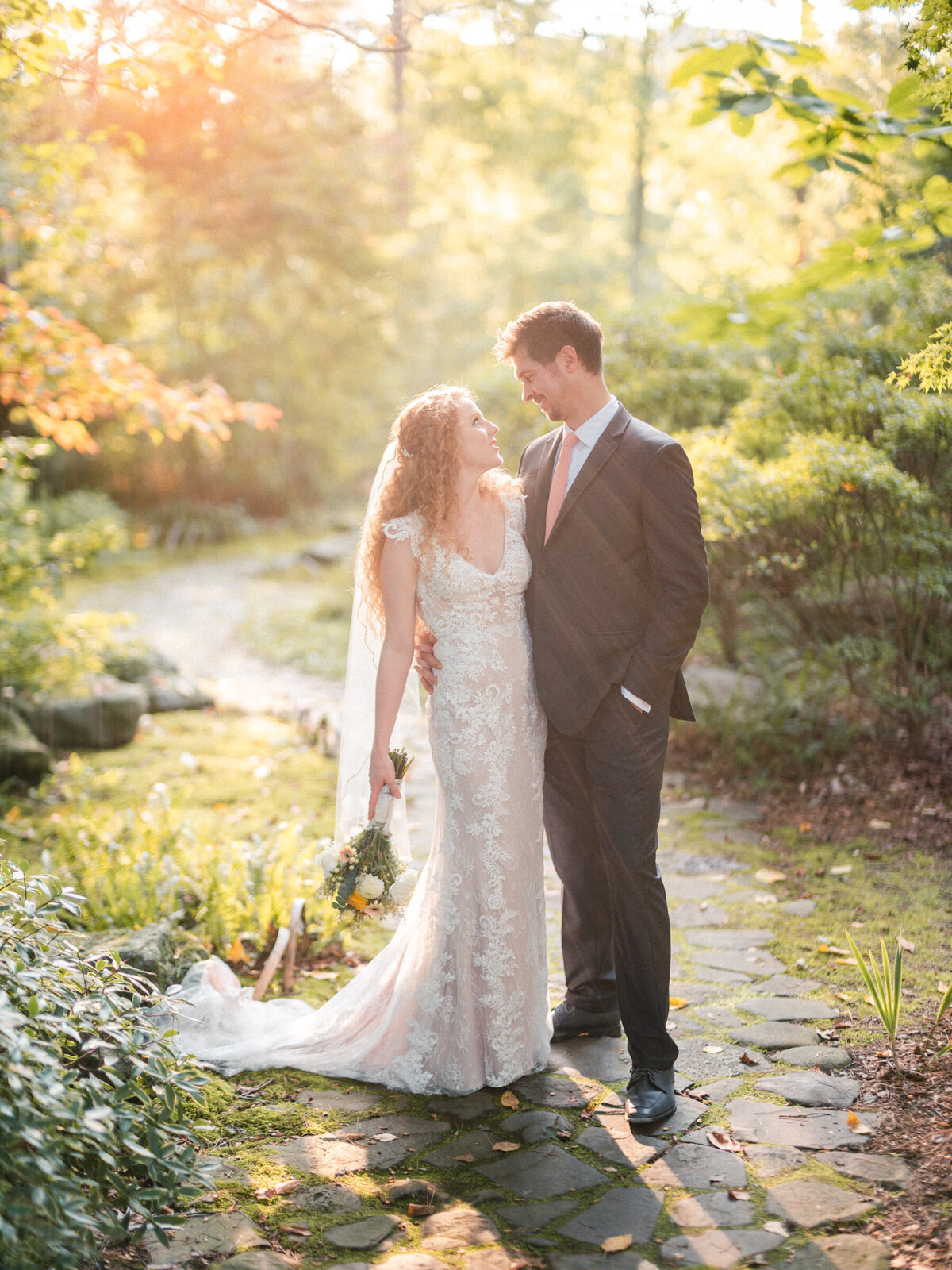 Bride and groom bathed in golden sunlight in a lush garden, sharing a tender moment on a cobblestone path