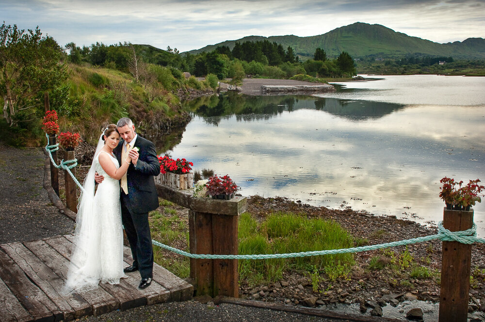 bride wearing an a-line style wedding dress with scooped back embracing her grey haired groom while standing on a wooden bridge overlooking Golden Cove, Sneem