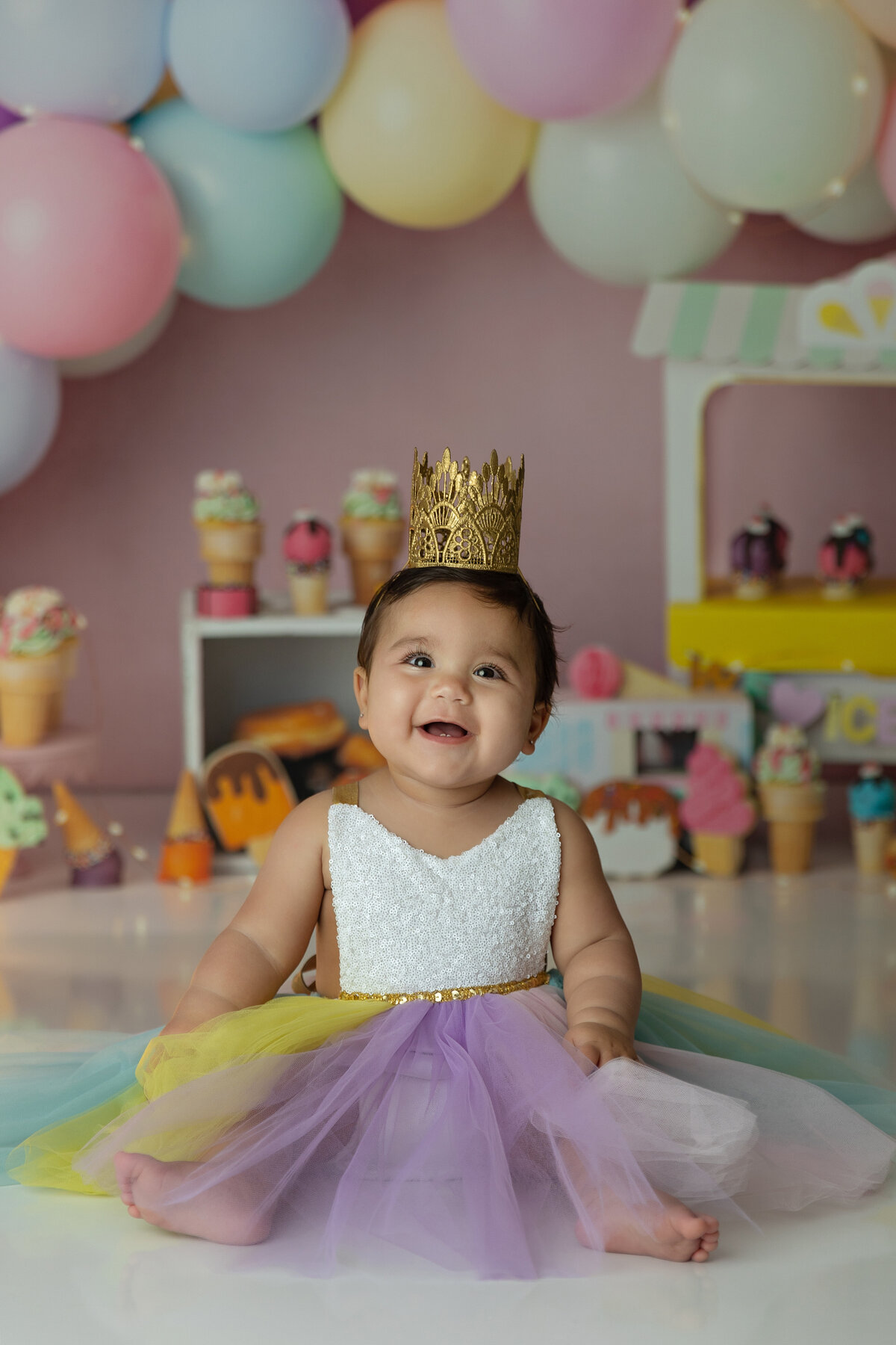 A happy toddler in a gold crown sits on the floor of a studio smiling for her first birthday