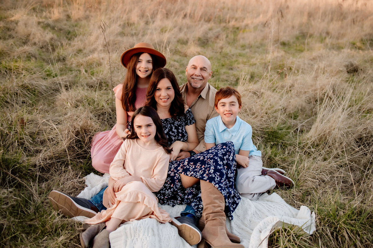 family of 5 snuggled on a blanket close together in a field at sunset