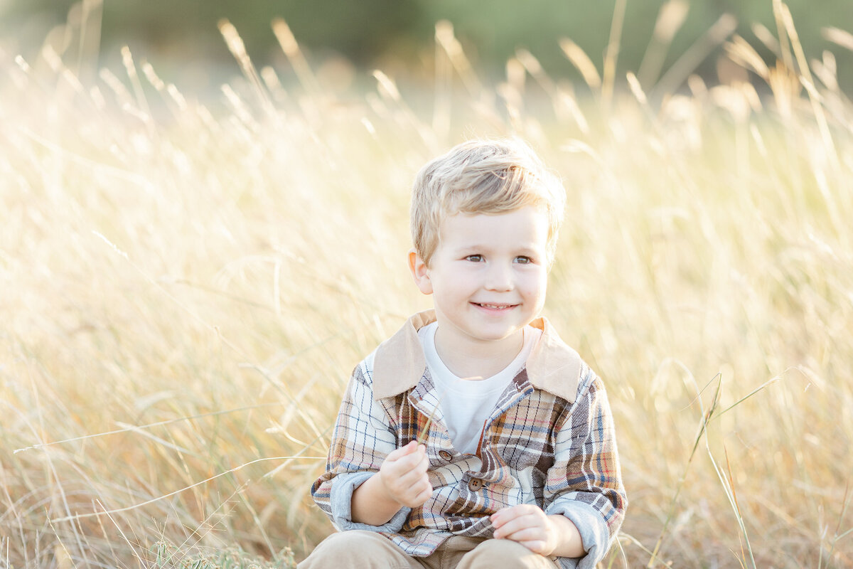 A little boy sitting in the grass as he looks off and smiles at his mother as they take family photos.