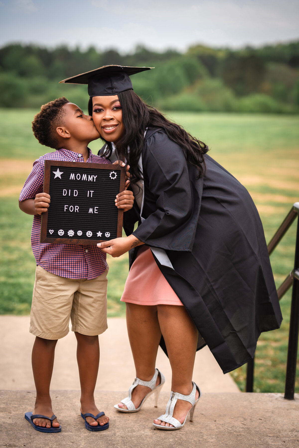 A mom in a graduation cap is doing a photoshoot with her young son and he is kissing her on the cheek.
