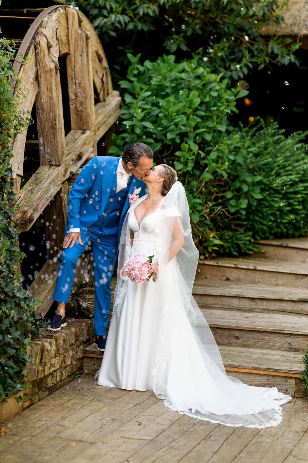 Bruid en bruidegom kussen elkaar liefdevol bij watermolen tijdens hun bruiloft, georganiseerd door weddingplanner