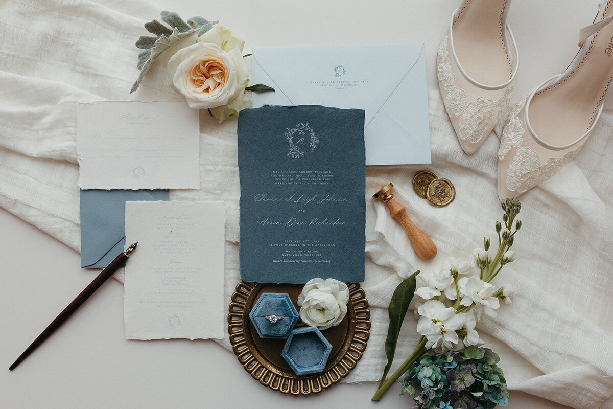 An assortment of blue and gray wedding stationery with white cursive next to a wedding ring, flowers and white wedding heels.
