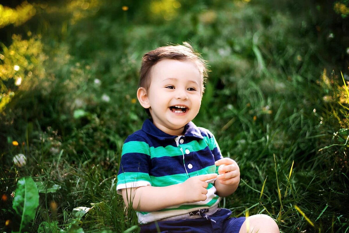 1 year old boy in green and blue laughing during outdoor milestone session in the park