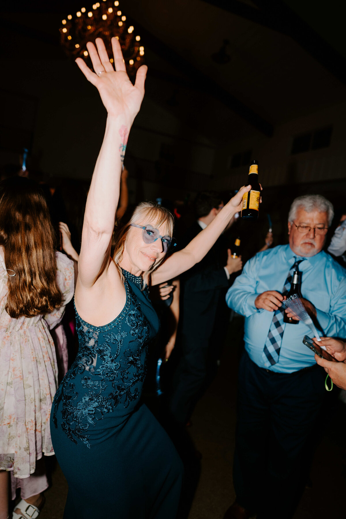 Woman in a teal dress joyfully dances with one arm raised, holding a drink in the other. People in the background, festive atmosphere, dim lighting.