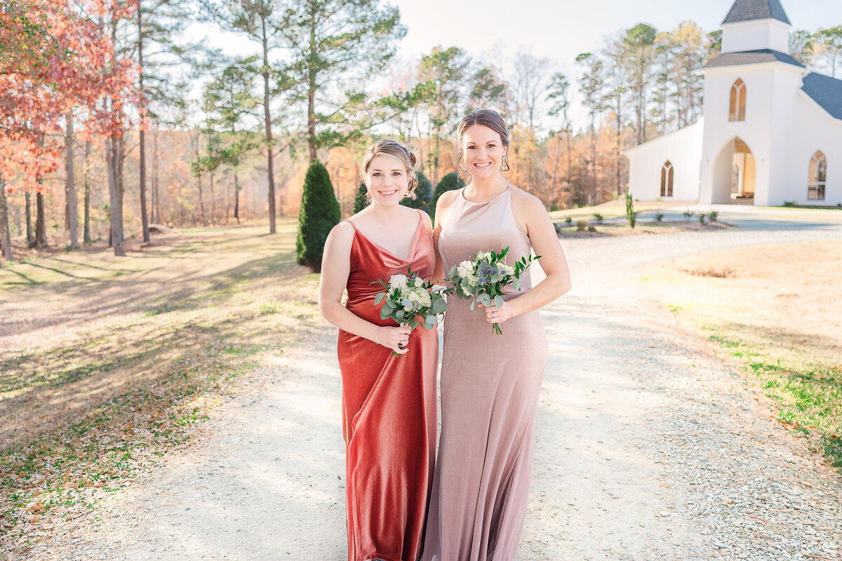 Two bridesmaids in warm dresses holding flower bouquets in the Raleigh countryside enjoying North Carolina wedding photos by JoLynn Photography