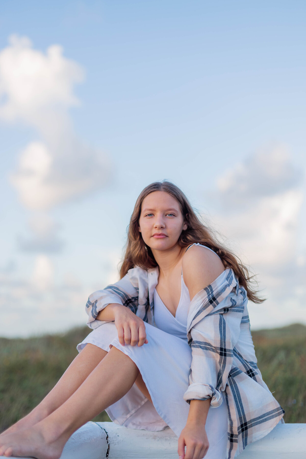 Senior girl sits on a fence in a white dress and flannel shirt with dunes in the background
