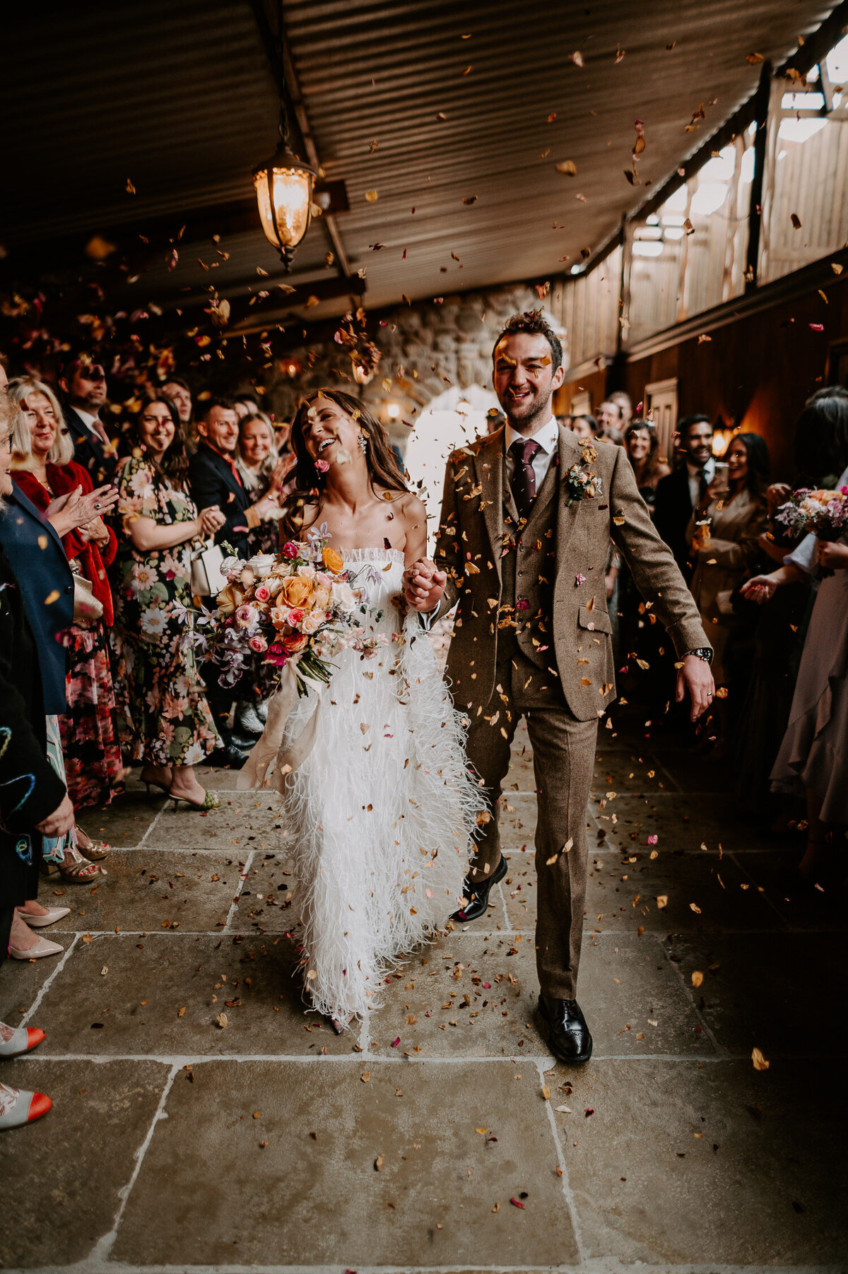 Confetti flys down on a bride and groom at The Willow Marsh Farm. The bride is also holding an orange bouquet and is wearing a fringed wedding dress.