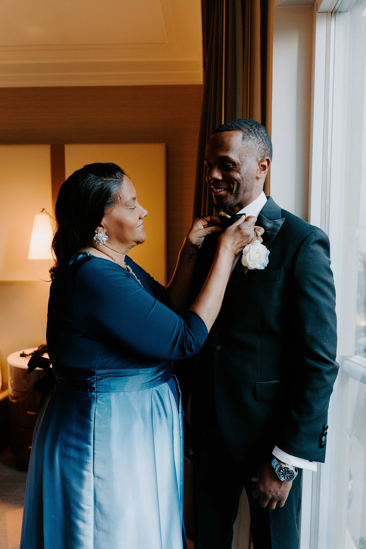 A woman in a blue dress adjusts the tie of a smiling man in a black suit by a window. The warm indoor lighting sets a joyful, intimate tone.