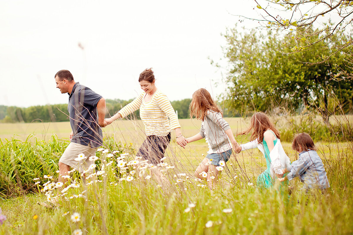 Family of five holding hands and walking through a tall field of grass, from big to little.