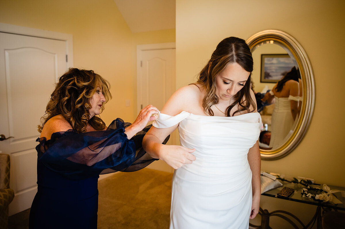 A woman in a strapless wedding dress getting assistance with her gown from another woman in a blue dress with a mirror reflecting the scene.