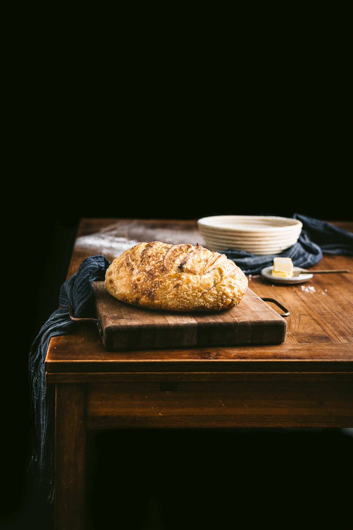 Oval shaped loaf of sourdough on wooden table with cutting board and flour dusted on table