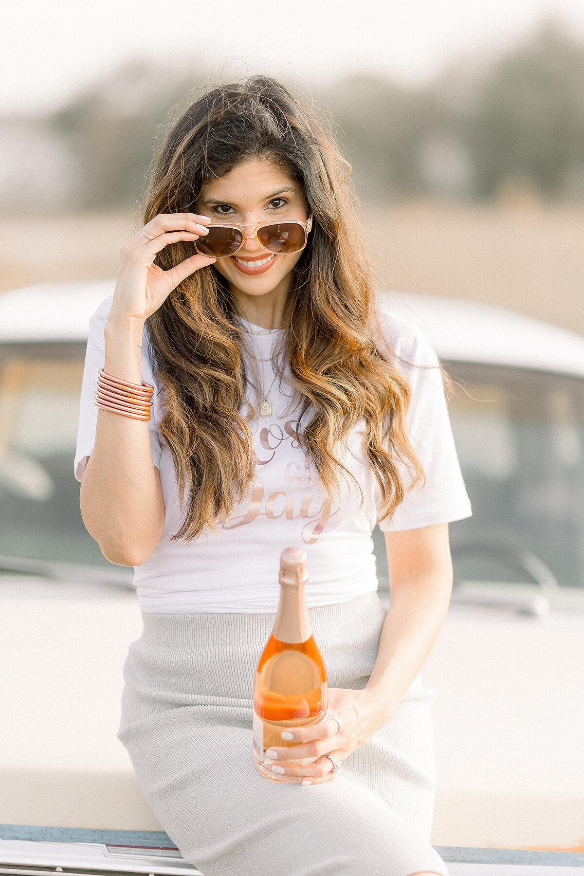 Photo of a professional DFW business owner sitting on the hood of a vintage car playfully posing for her headshot photos and pulling her glasses down from her face.